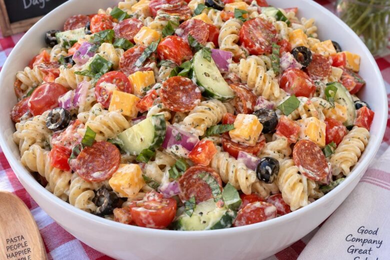 Photo of Creamy Italian Pasta Salad with Pepperoni and Fresh Veggies (creamy italian pasta salad): A large white serving bowl of creamy italian pasta salad made with rotini, halved cherry tomatoes, cucumber slices, diced red bell pepper, red onion, mini pepperoni, cheddar cubes, black olives, and fresh basil, photographed in bright natural light on a picnic table