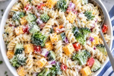 Photo of Creamy Ranch Pasta Salad with Cheddar and Crunchy Veggies (creamy ranch pasta salad): A bright overhead food photograph of creamy ranch pasta salad in a large white serving bowl, with rotini coated in creamy herb dressing, cheddar cheese cubes, broccoli, diced cucumber, red bell pepper, and red onion clearly visible, a serving spoon resting in the bowl