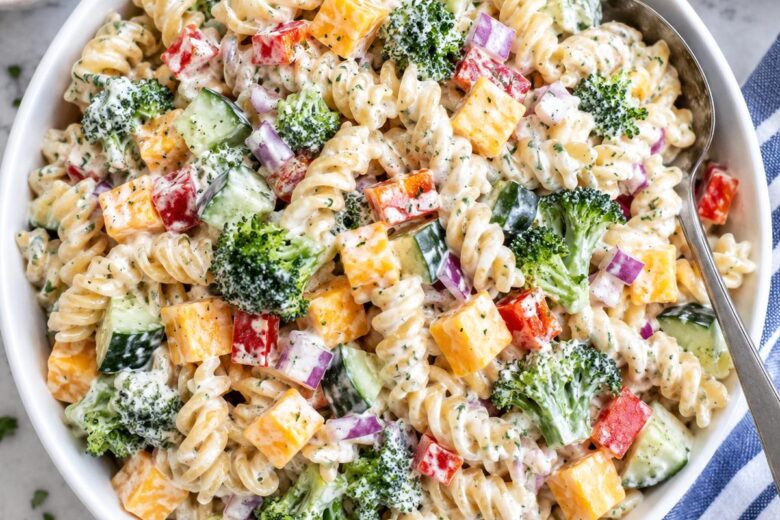 Photo of Creamy Ranch Pasta Salad with Cheddar and Crunchy Veggies (creamy ranch pasta salad): A bright overhead food photograph of creamy ranch pasta salad in a large white serving bowl, with rotini coated in creamy herb dressing, cheddar cheese cubes, broccoli, diced cucumber, red bell pepper, and red onion clearly visible, a serving spoon resting in the bowl