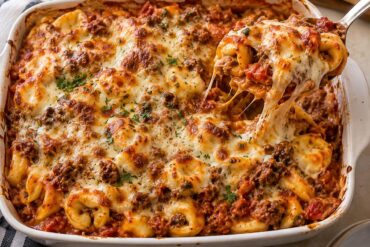 Photo of Ground Beef Pasta Casserole Recipes: Cheesy Tortellini Bake (ground beef pasta casserole recipes): Overhead view of a baked ground beef pasta casserole recipe made with cheese tortellini, marinara, browned beef, and a bubbling golden mozzarella-cheddar topping in a white 9-by-13-inch baking dish, with a serving spoon lifting a portion