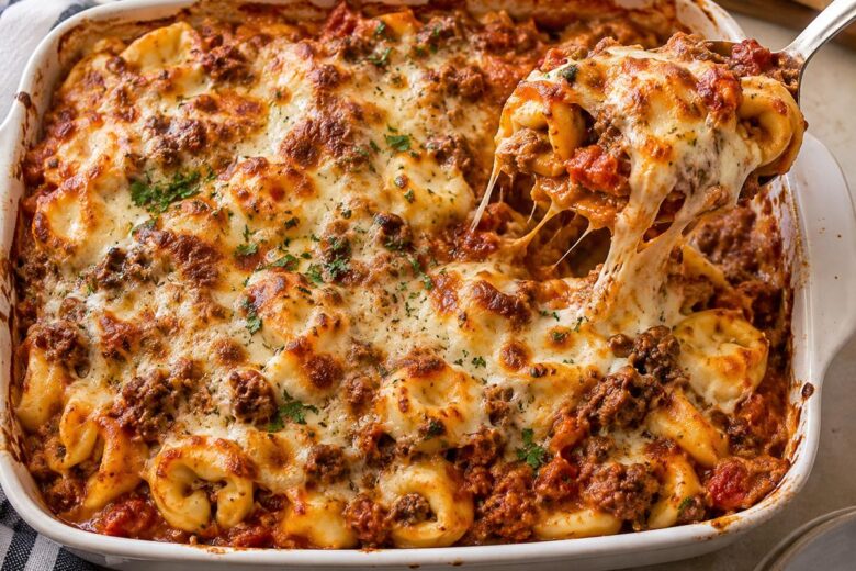 Ground Beef Pasta Casserole Recipes: Cheesy Tortellini Bake 15 Photo of Ground Beef Pasta Casserole Recipes: Cheesy Tortellini Bake (ground beef pasta casserole recipes): Overhead view of a baked ground beef pasta casserole recipe made with cheese tortellini, marinara, browned beef, and a bubbling golden mozzarella-cheddar topping in a white 9-by-13-inch baking dish, with a serving spoon lifting a portion