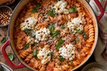Photo of Creamy Tomato One Pot Pasta Recipes with Ricotta (one pot pasta recipes): A rustic Dutch oven filled with creamy tomato one pot pasta recipes, short fusilli coated in glossy red sauce with dollops of ricotta, torn basil, Parmesan, and red chili flakes, photographed overhead on a warm kitchen table