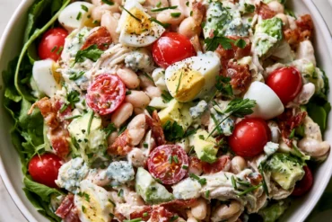 Photo of Creamy High Fiber Chicken Salad with White Beans and Avocado (high fiber chicken salad): A bright overhead food photograph of high fiber chicken salad in a wide white serving bowl, showing shredded chicken, white beans, diced avocado, halved grape tomatoes, chopped hard-boiled eggs, crisp bacon, blue cheese, chives, and parsley in creamy dressing, with leafy greens around the bowl
