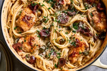 Photo of Creamy One Pot Mediterranean Pasta with Chicken, Lemon, and Olives (one pot mediterranean pasta): A warm overhead food photograph of creamy one pot Mediterranean pasta with chicken in a wide Dutch oven, showing long fettuccine coated in lemon cream sauce, golden chicken pieces, halved Kalamata olives, capers, grated Parmesan, and chopped parsley
